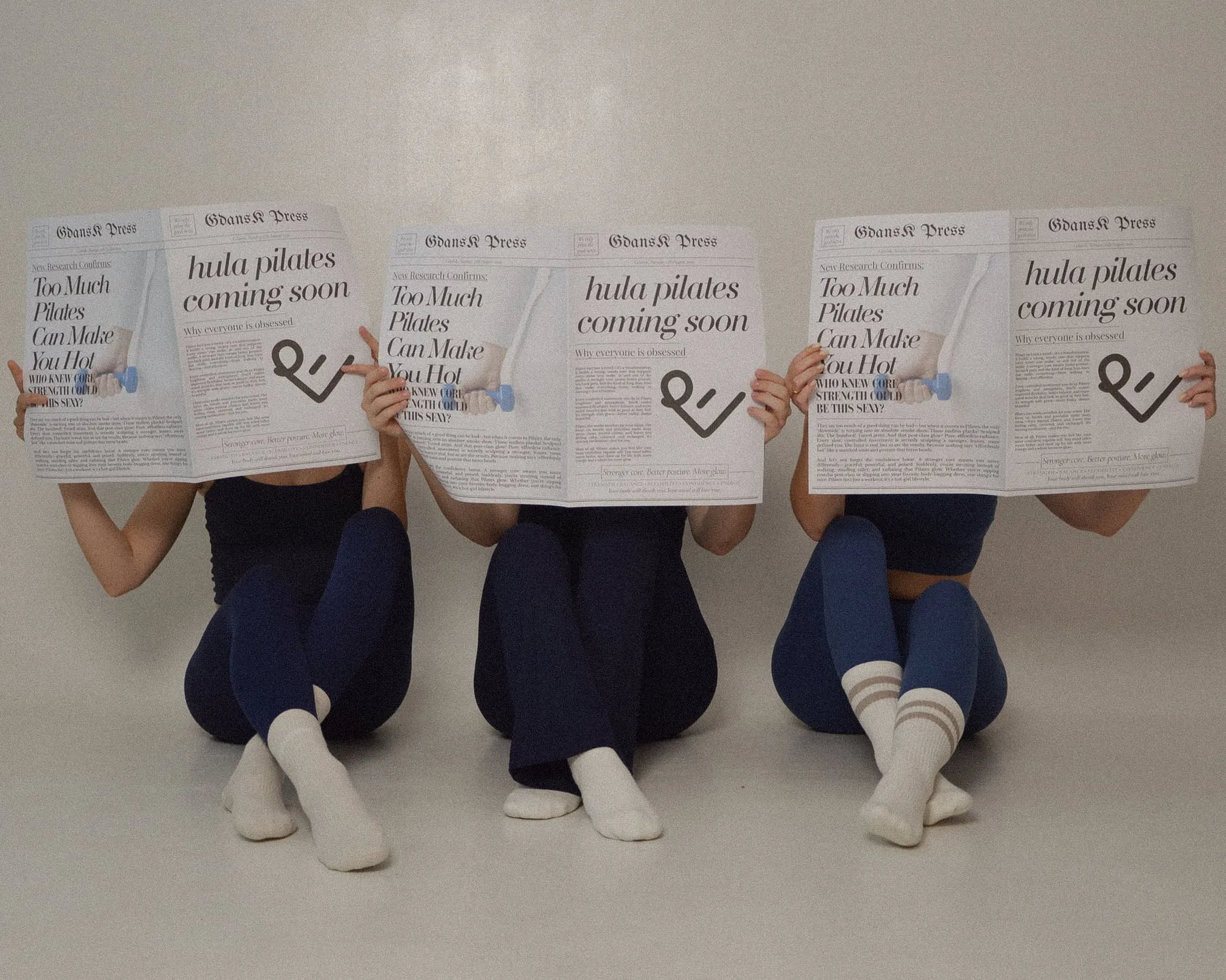 Trzy osoby siedzą za gazetami z nagłówkiem „hula pilates coming soon”